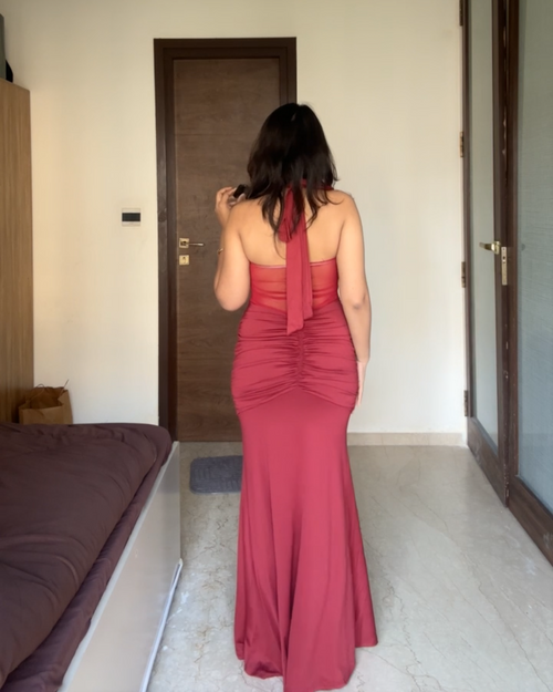 A woman viewed from behind wearing a floor-length dusty rose pink halter gown in a bedroom setting.