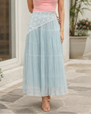 A woman modeling a sleeveless halter-neck maxi dress with a pink cowl neckline and a tiered light blue skirt with white floral embroidery, standing on a stone walkway in front of a white building.